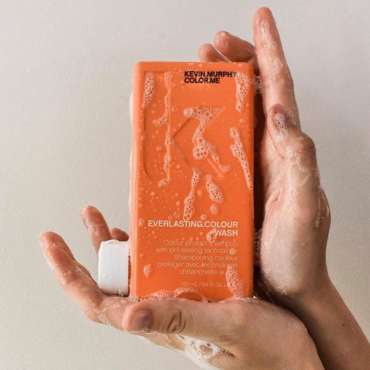 Hands holding a bottle of Kevin Murphy Everlasting Colour Wash shampoo against a neutral background, showcasing the product's details and foamy texture
